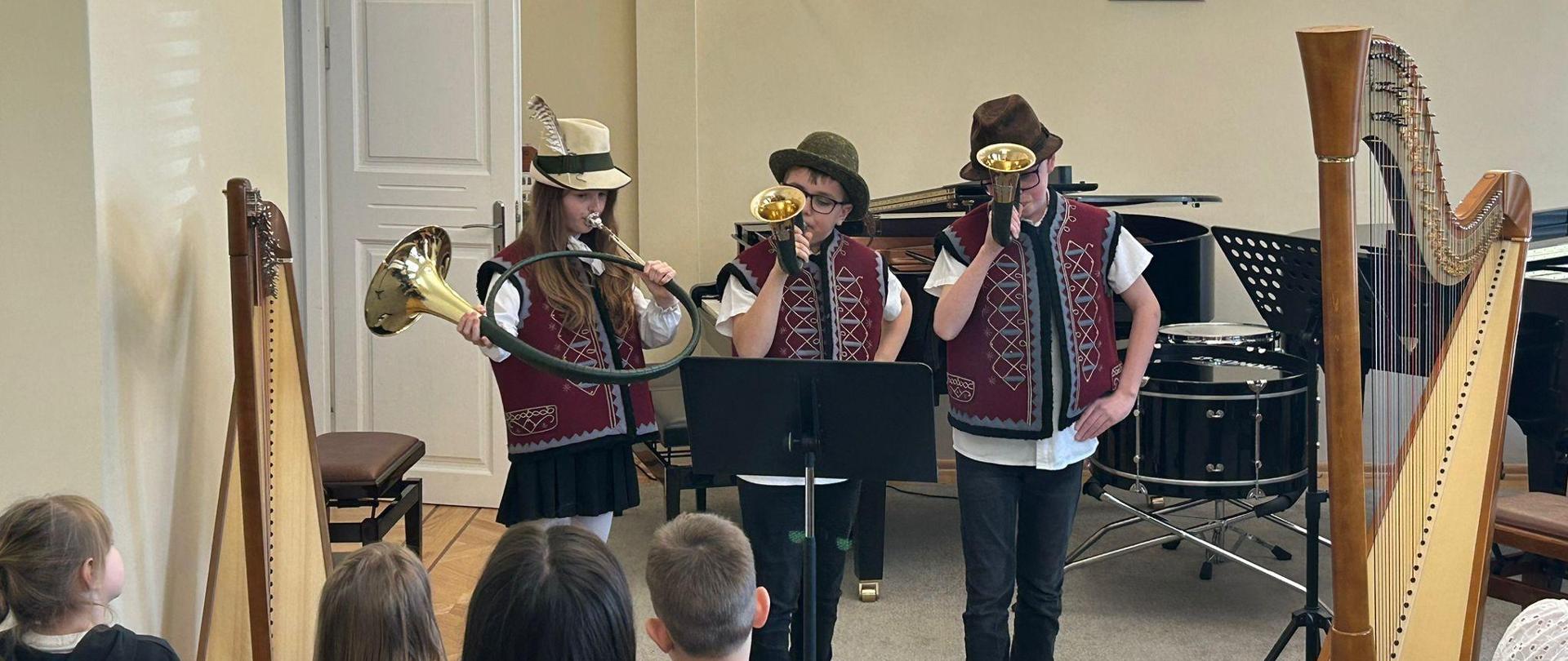 Troje uczniów grających na rogach myśliwskich w auli szkolnej. Tyłem siedzi publiczność.