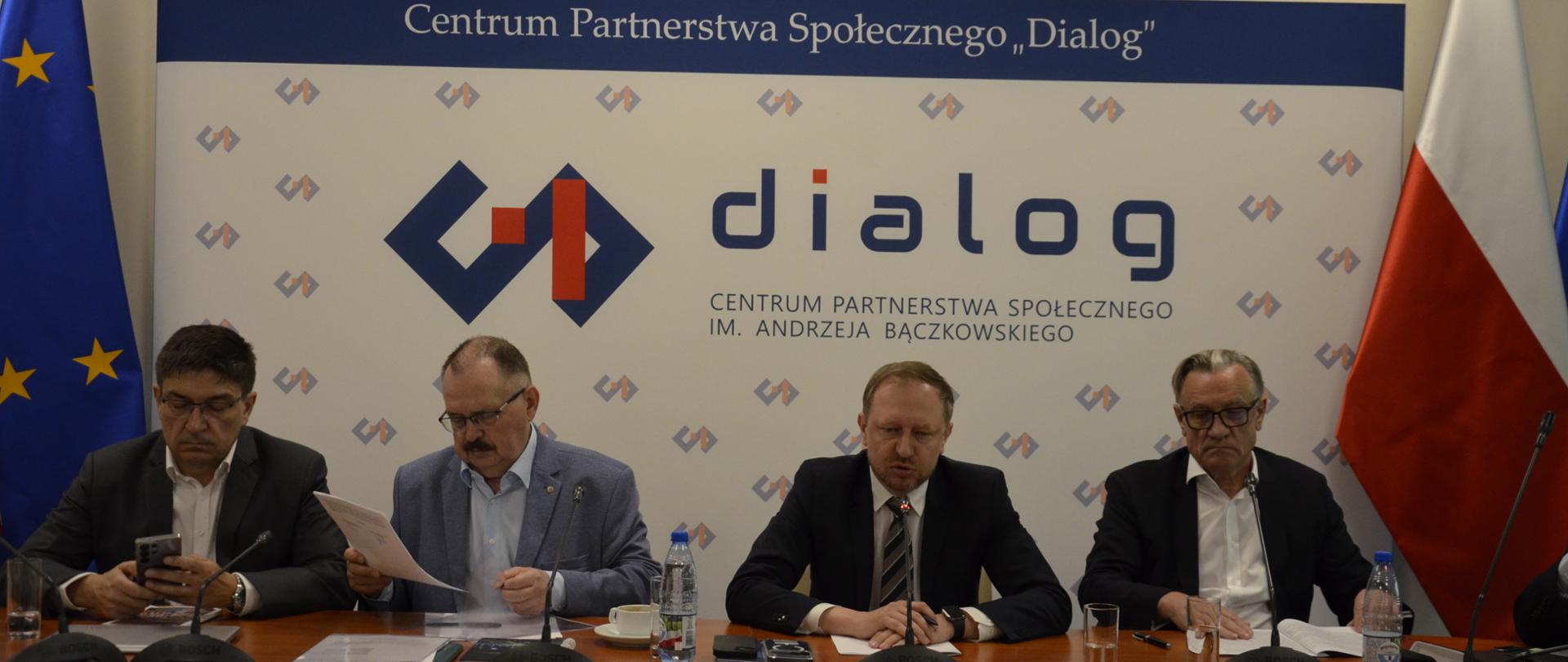 Na zdjęciu widnieje stół prezydialny za którym siedzą: Pan Wojciech Wrochna, Sekretarz Stanu w Ministerstwie Energii; Pan Wojciech Ilnicki, Współprzewodniczący Zespołu Trójstronnego ds. Branży Węgla Brunatnego ze strony związków zawodowych; Pan Jerzy Wiertelak, Współprzewodniczący Zespołu Trójstronnego ds. Branży Energetycznej ze strony związków zawodowych oraz pan Jacek Kaczorowski, Współprzewodniczący Zespołu Trójstronnego ds. Branży Węgla Brunatnego ze strony pracodawców. W tle znajduje się baner z napisem Centrum Partnerstwa Społecznego „Dialog” im. Andrzeja Bączkowskiego. Po lewej stronie banneru znajduje się flaga UE, po prawej flaga Polski. Na górze znajduje się Godło Polski.