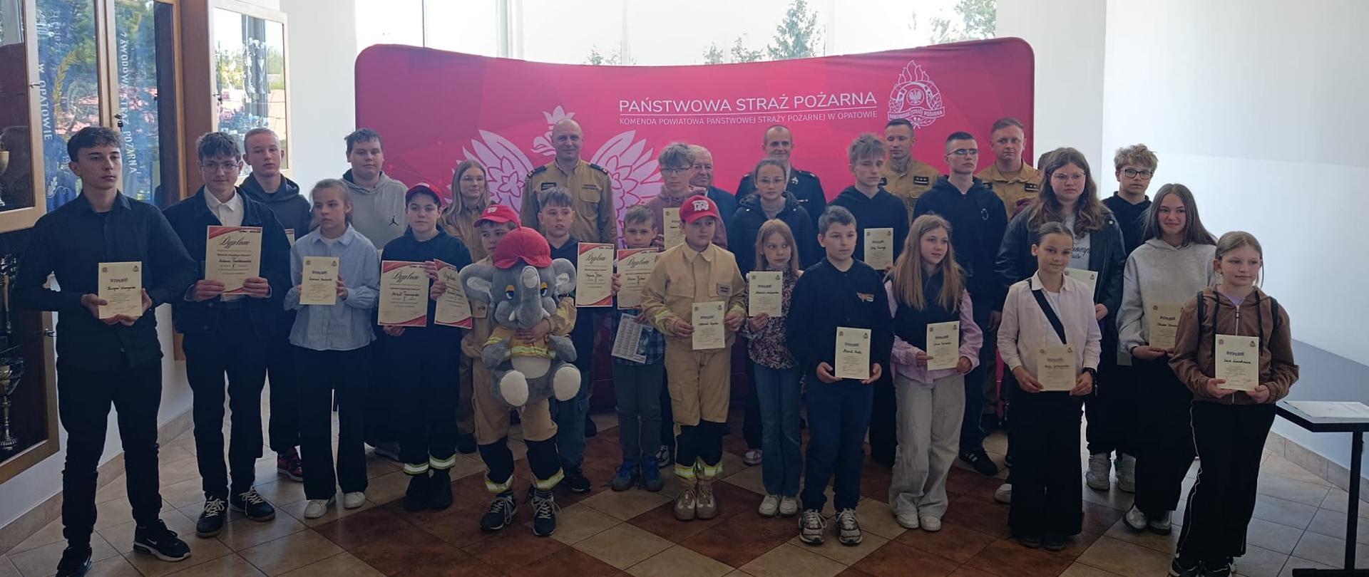 Grupa dzieci, młodzieży i dorosłych pozuje do pamiątkowego zdjęcia w jasnej sali. Uczestnicy trzymają dyplomy, a część osób ma na sobie stroje strażackie. W tle znajduje się różowy baner z napisem „Państwowa Straż Pożarna” oraz gabloty z eksponatami.