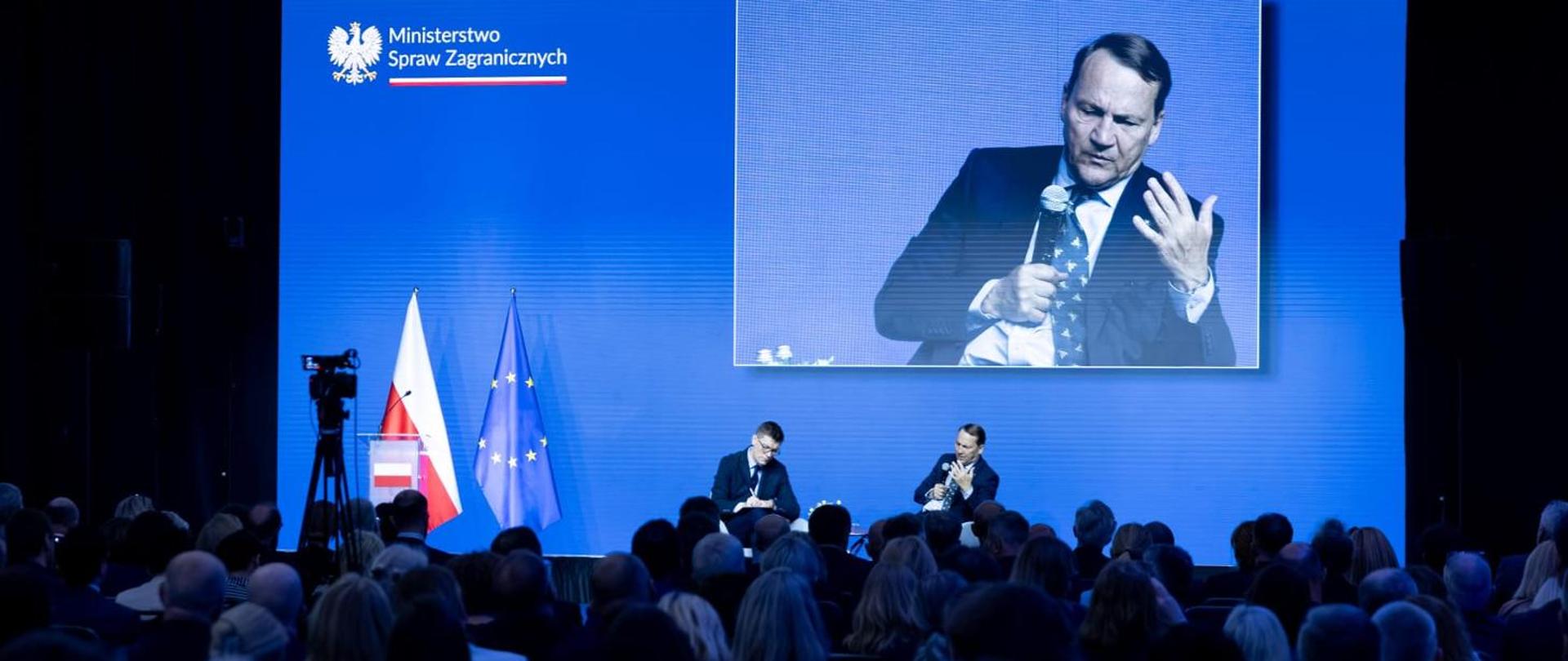 The photo shows two men in suits on stage. They are speaking into microphones. A blue light shines on them. A group of several dozen people is listening to them. 