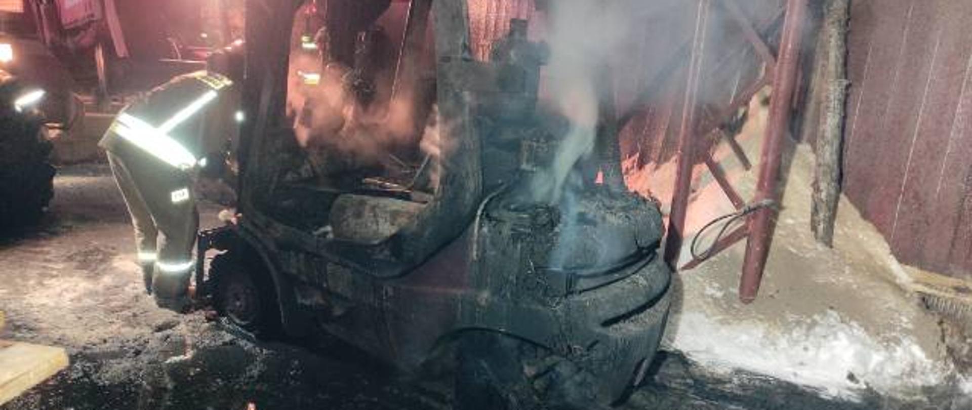 Zdjęcie przedstawia spalony wózek widłowy w skutek pożaru. Z maszyny unosi się jeszcze dym. Z lewej strony wózka stoi strażak ubrany w żółte ubranie specjalne. W tle widać ścianę budynku. Zdjęcie wykonane w porze nocnej. 