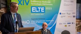 Wiceminister Murdzek stoi za drewnianą mównicą i mówi do mikrofonu, obok mównicy przykryty czerwonym materiałem stół za którym siedzi mężczyzna w garniturze i okularach, za nimi rozwijany baner z napisem XIV konferencja naukowa technologia elektronowa ELTE 2023.
