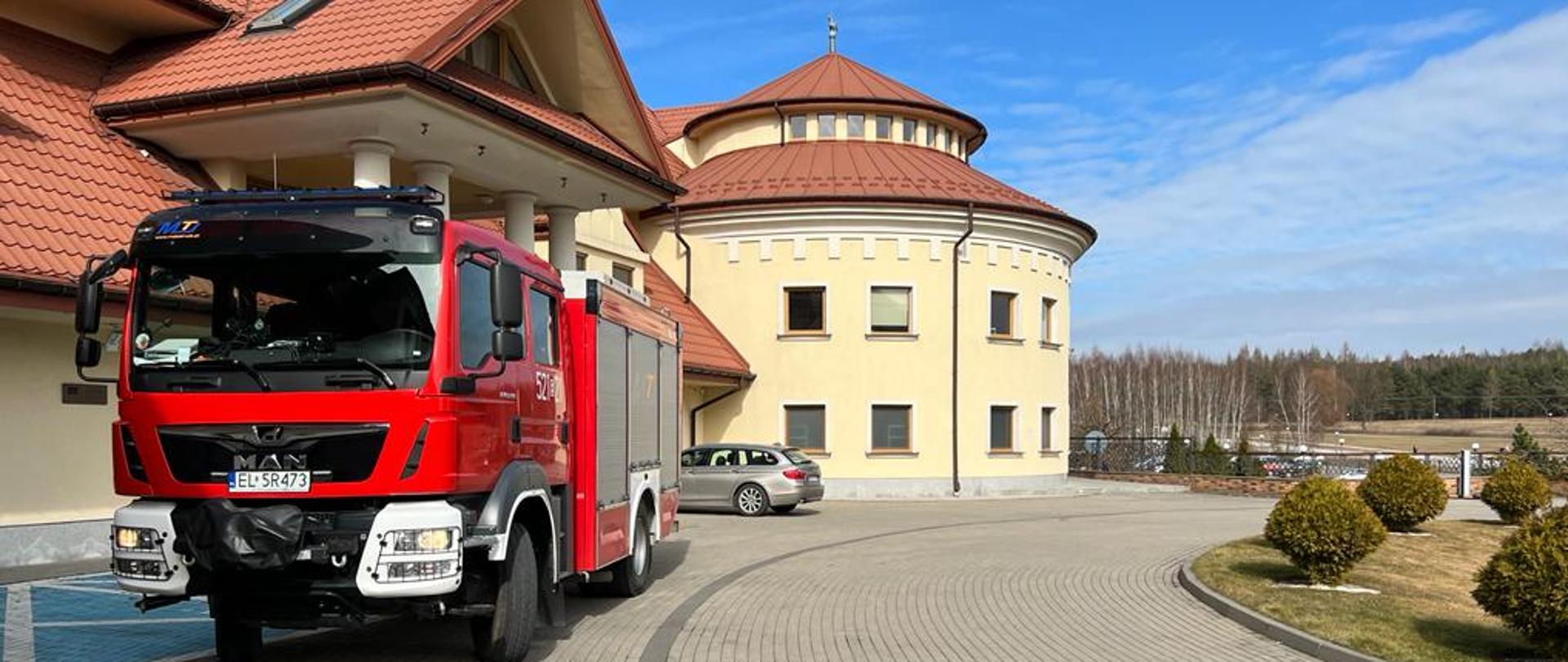 Przed budynkiem hotelu, na podjeździe z koski betonowej stoi samochód strażacki. Hotel ma żółte ściany i dach w kolorze cegłówki. Na prawo, w oddali widać lasy.