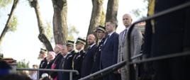 Widok z boku na podwyższenie, na którym stoi minister Czarnek i strażacy w czarnych galowych mundurach.