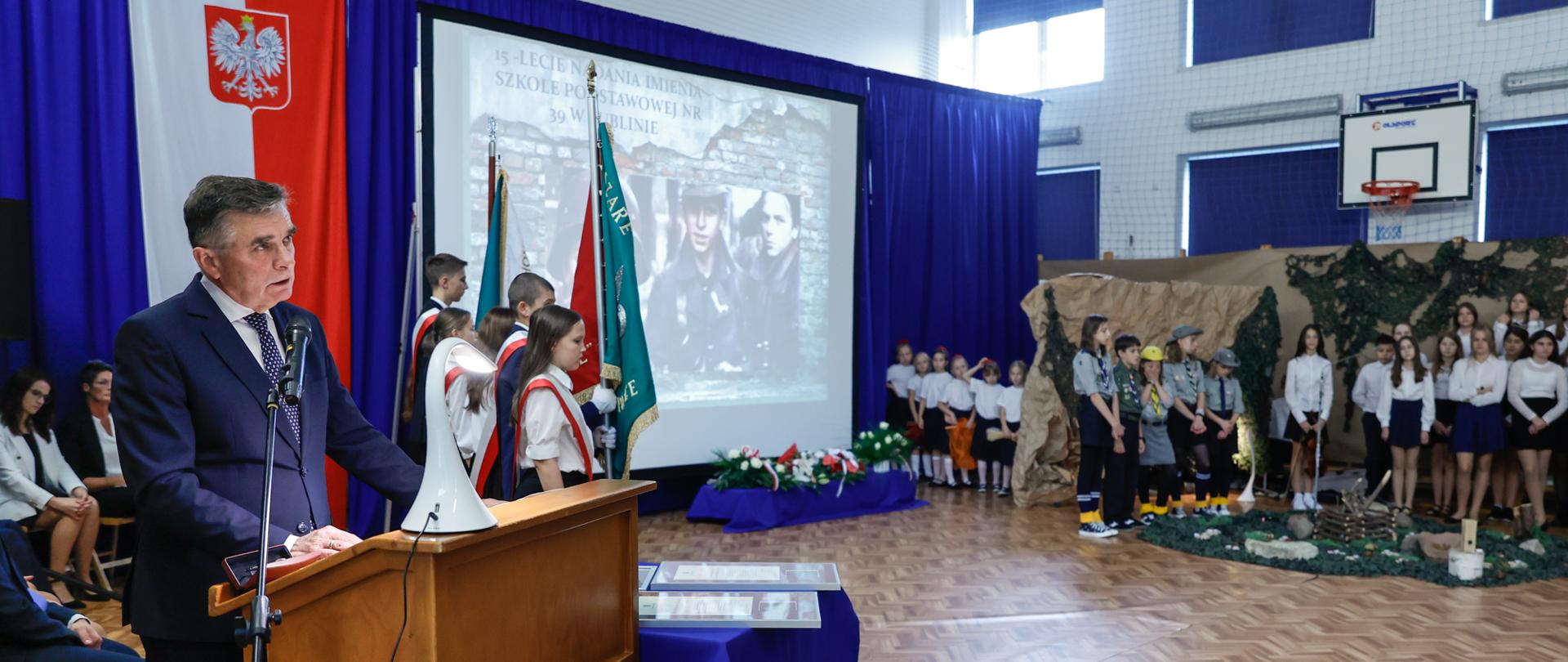 Wojewoda stoi przy mównicy, przemawia. Widać dzieci ze sztandarem, dyplomy uznania na stoliku, dzieci-harcerzy.