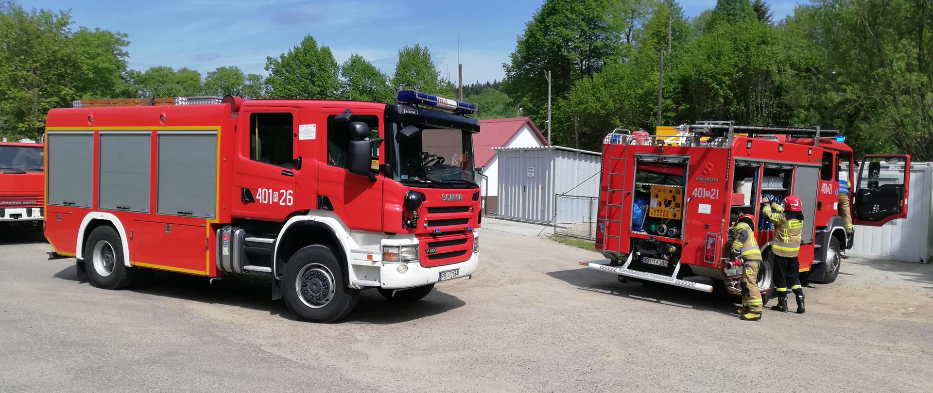 Na zdjęciu widać dwa pojazdy pożarnicze z JRG Ustrzyki Dolne jeden z nich to scania który stoi bokiem, drugi to Man stojący tyłem z otwartymi skrytkami. Przy Manie jest dwóch strażaków w ubraniach specjalnych którzy wyciągają sprzęt ze skrytek. W tle znajdują się blaszane garaże.