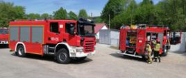 Na zdjęciu widać dwa pojazdy pożarnicze z JRG Ustrzyki Dolne jeden z nich to scania który stoi bokiem, drugi to Man stojący tyłem z otwartymi skrytkami. Przy Manie jest dwóch strażaków w ubraniach specjalnych którzy wyciągają sprzęt ze skrytek. W tle znajdują się blaszane garaże.
