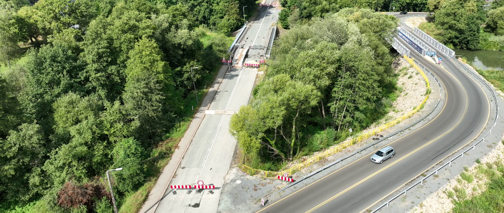 Zdjęcie przedstawia widok z lotu ptaka na fragment drogi w otoczeniu zieleni. Po lewej stronie znajduje się zamknięty odcinek jezdni, zabezpieczony barierami w biało-czerwone pasy.