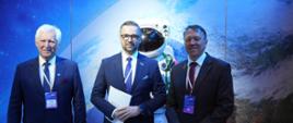 Na tle wielkiego zdjęcia przedstawiającego astronautę na tle Ziemi stoi minister Kulasek pomiędzy dwoma mężczyznami w garniturach.