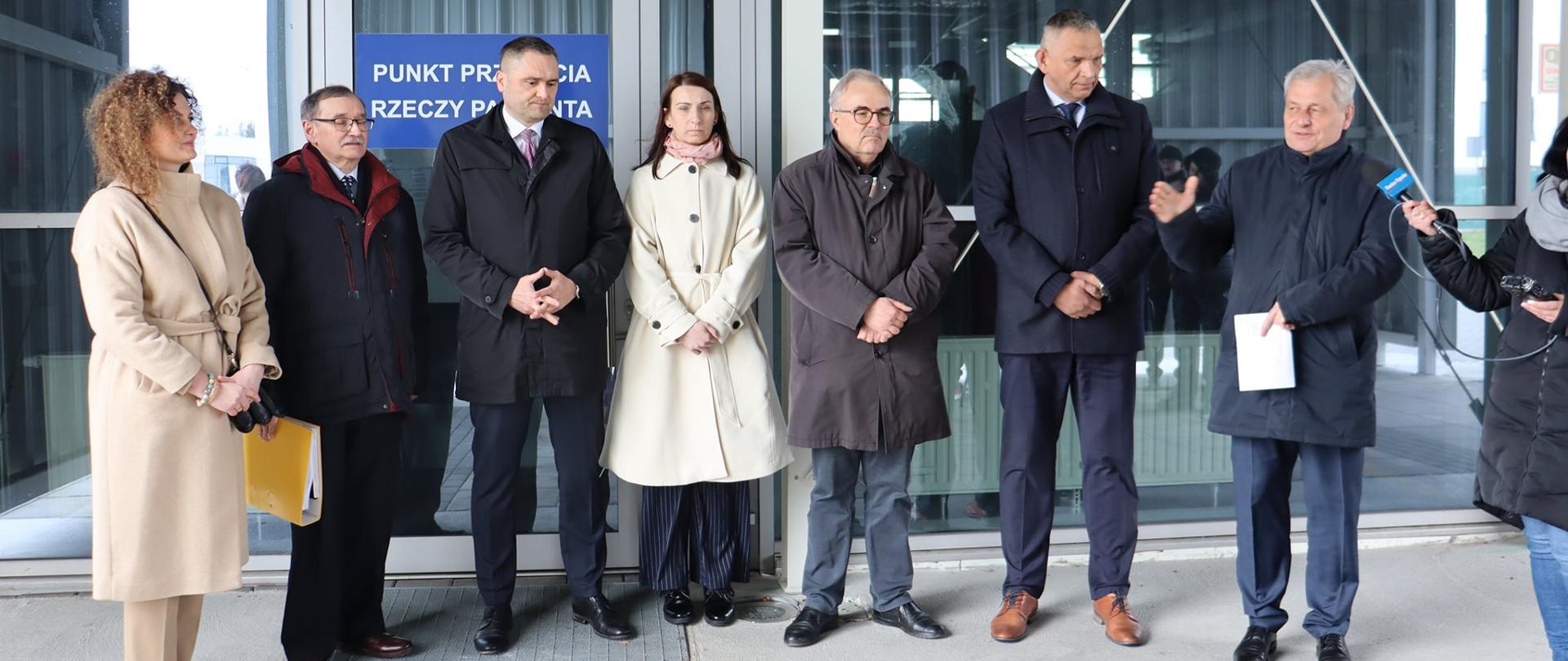 Na zdjęciu (od lewej) Wojewoda opolski, dyrekcja USK w Opolu, kierownik szpitala tymczasowego oraz pracownicy OUW zaangażowani w prace obiektu . Łącznie 7 osób. Po prawej stornie kadru widać dziennikarza trzymającego mikrofon. W tle szyba.