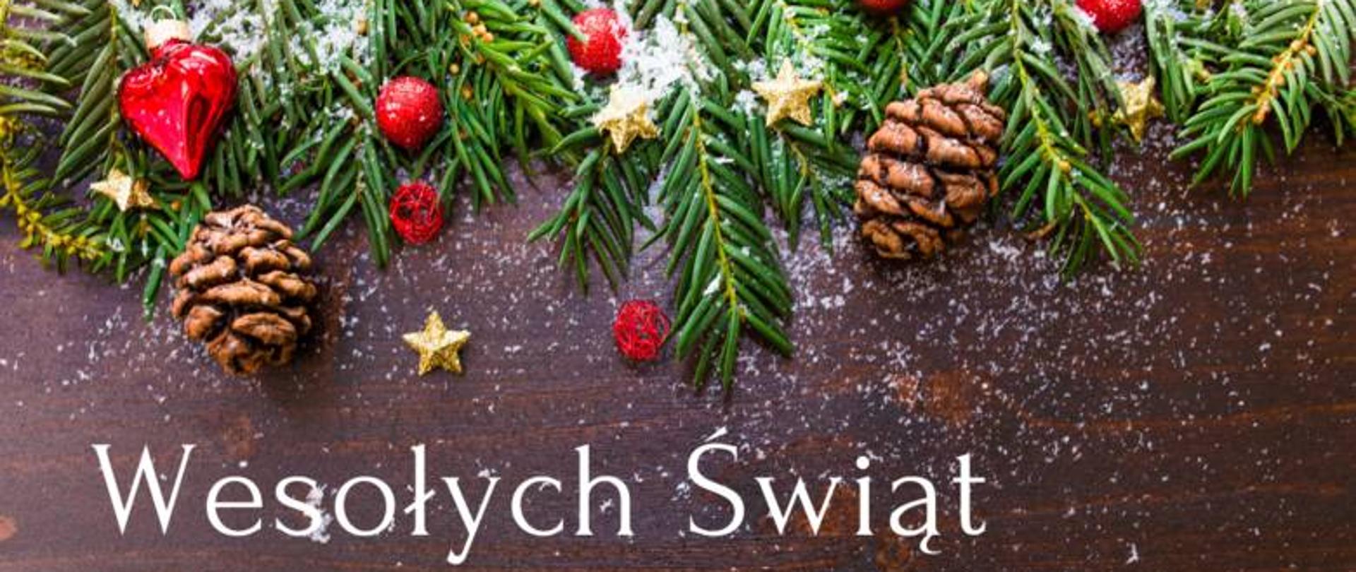Karta okolicznościowa z okazji Świąt Bożego Narodzenia. Na drewnianym stole w kolorze ciemnobrązowym ułożone zielone gałązki świerka. Od góry zdjęcia znajdują się gałązki skierowane ku dołowi. Pomiędzy gałązkami ułożono dla ozdoby czerwone jagody, czerwoną bombkę w kształcie serca oraz dwie szyszki. Na dole napis "Wesołych Świąt" 