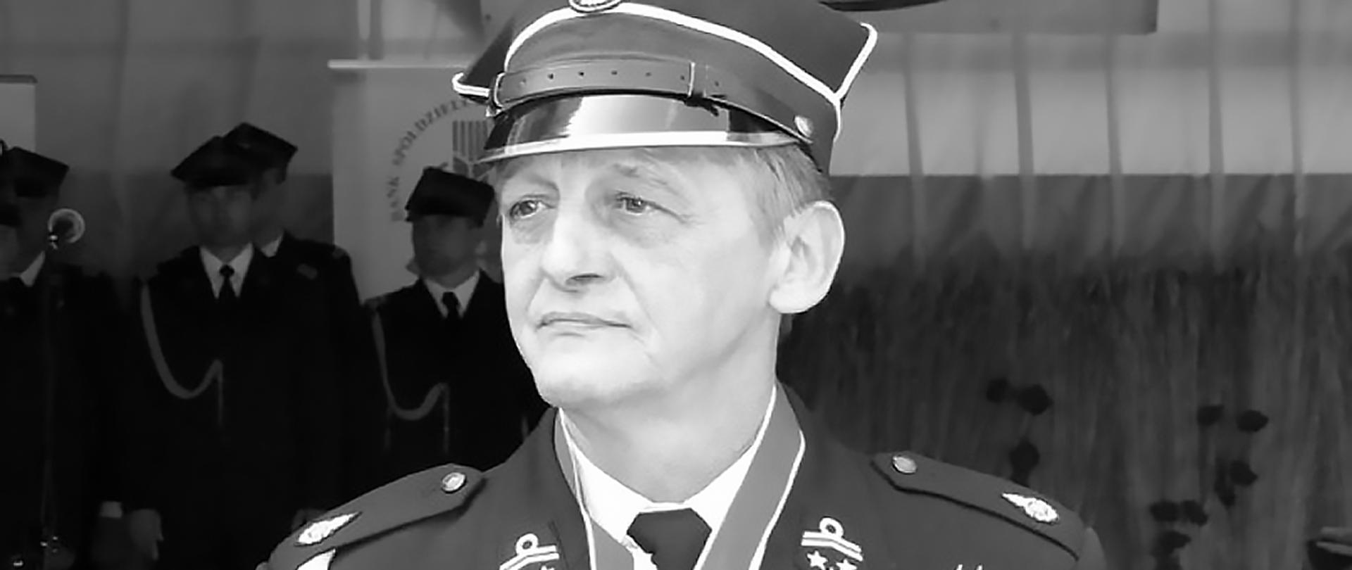 Zdjęcie przedstawia Prezesa OSP Ratownictwo Wodne w Łowiczu, ubrany jest w mundur galowy ze sznurem i czapką rogatywką na głowie, na szyi ma szarfę na której zawieszono odznaczenie, w tle funkcjonariusze Jednostki Ratowniczo-Gaśniczej komendy powiatowej PSP w Łowiczu ubrani w mundury galowe ze sznurem i rogatywka na głowie. Zdjęcie wykonano w technice czarno-białej.
