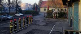 Zmiana służbowa w umundurowaniu specjalnym stoi w szeregu na placu JRG. W tle strażacy salutują pod masztem.