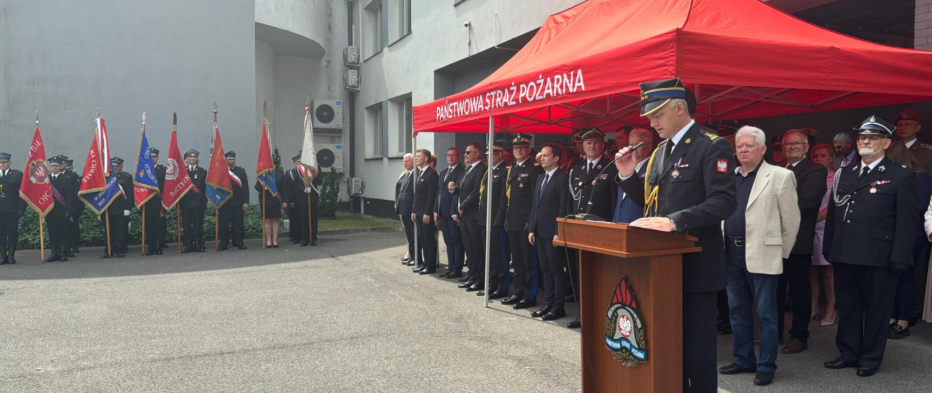 Strażak w mundurze galowym przemawia z mównicy na tle czerwonego namiotu, pod którym stoją zgromadzeni goście. W oddali, pod dużym jasnym budynkiem z dużymi oknami stoją w szeregu strażacy ze sztandarami jednostek straży pożarnych. 