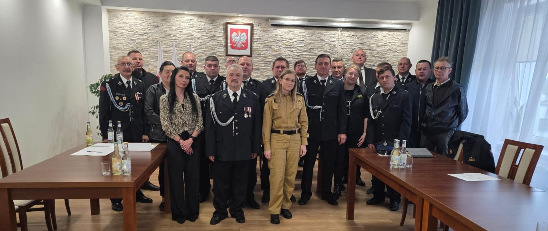 Grupa kilkunastu osób stoi do wspólnego zdjęcia w jasnej sali konferencyjnej. Większość uczestników ma na sobie ciemne mundury strażackie lub galowe, a na środku stoi młoda kobieta w beżowym mundurze. W tle widoczna jest ściana z jasnego kamienia oraz godło Polski zawieszone centralnie nad grupą. Po bokach znajdują się drewniane stoły z butelkami i dokumentami, a po prawej stronie pod sufitem zamontowany jest projektor.