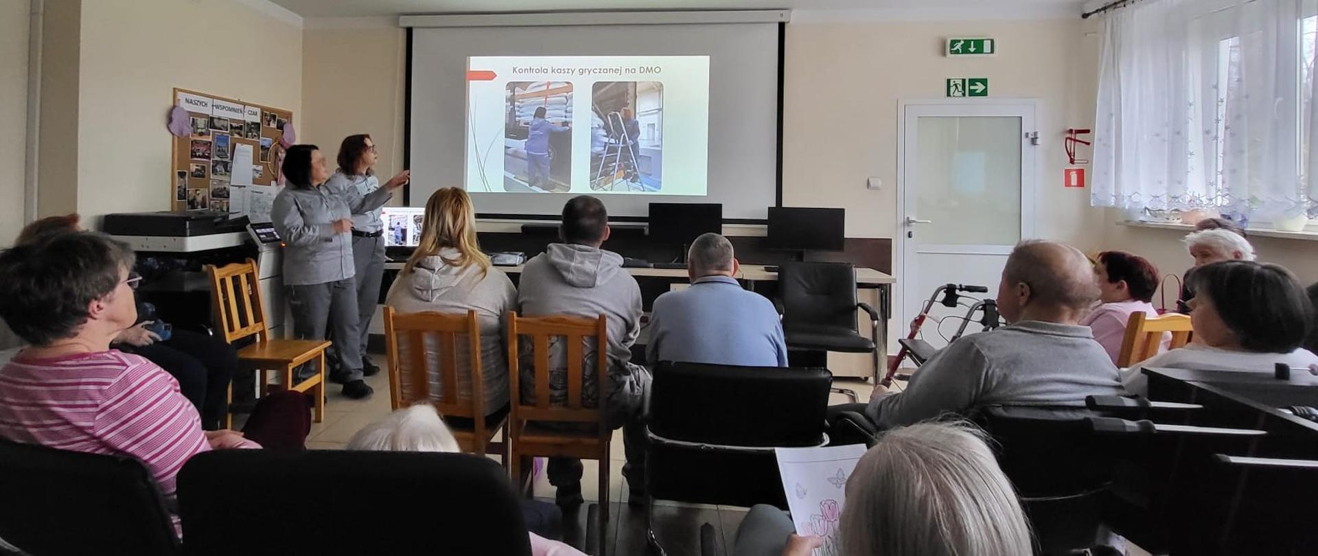  Od lewej: dwie kobiety w mundurach stoją i omawiają slajd wyświetlony na rzutniku w tle siedzący uczestnicy spotkania
