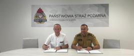 Komendant Miejski oraz przedstawiciel lasow państwowych. Siedzą przy stole i przymierzają się do podpisania porozumienia. W tle Napis Państwowa Straż Pożarna oraz logo PSP.