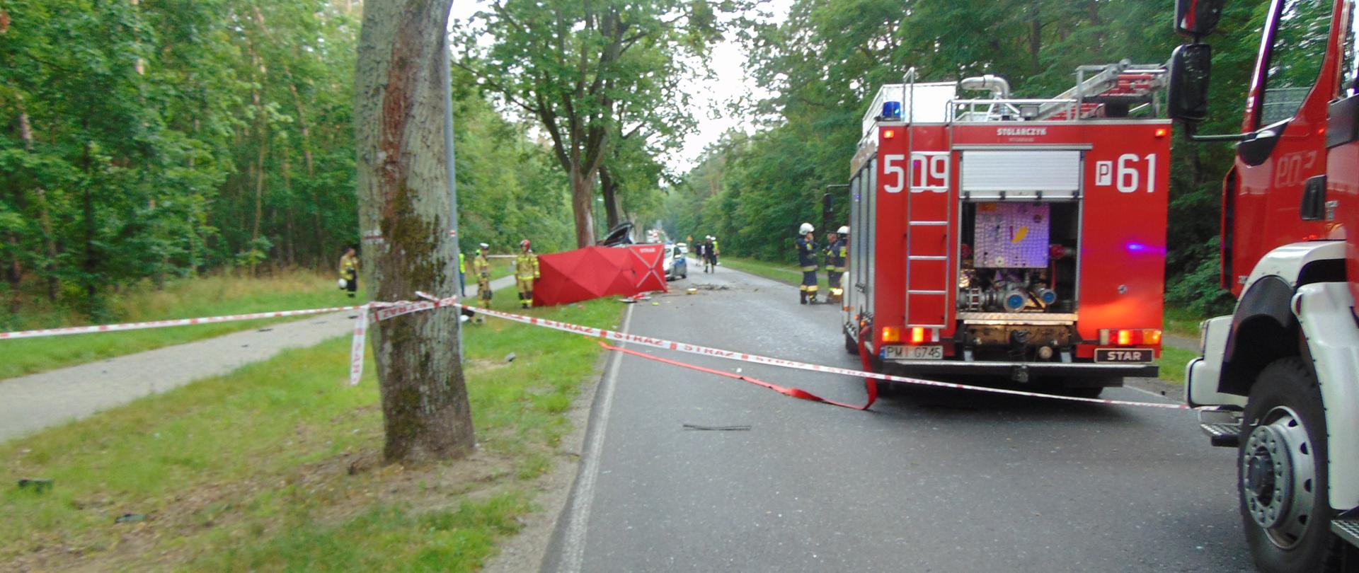 Na drodze stoi samochód straży pożarnej. W oddali widać parawan zasłaniający miejsce wypadku.