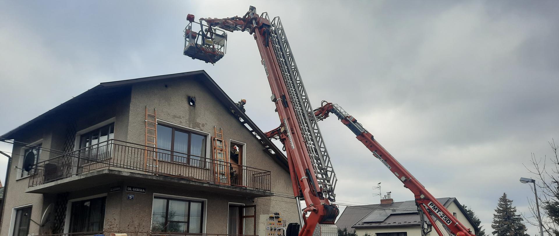 Zdjęcie przedstawia budynek mieszkalny w którym paliło się poddasze/ Na zdjęciu widać rozłożone dwa podnośniki hydrauliczne z drabiną. Ramienia są rozłożone i przystawione do dachu budynku. Na dachu widać strażaka ubranego w ubranie specjalne i hełm oraz aparat ochrony dróg oddechowych. Który usuwa nadpalone części dachu. Na balkonie widać przystawione dwie drabiny przystawne. Drzwi na balkony są otwarte. Po prawej stronie widać drugi budynek mieszkalny.