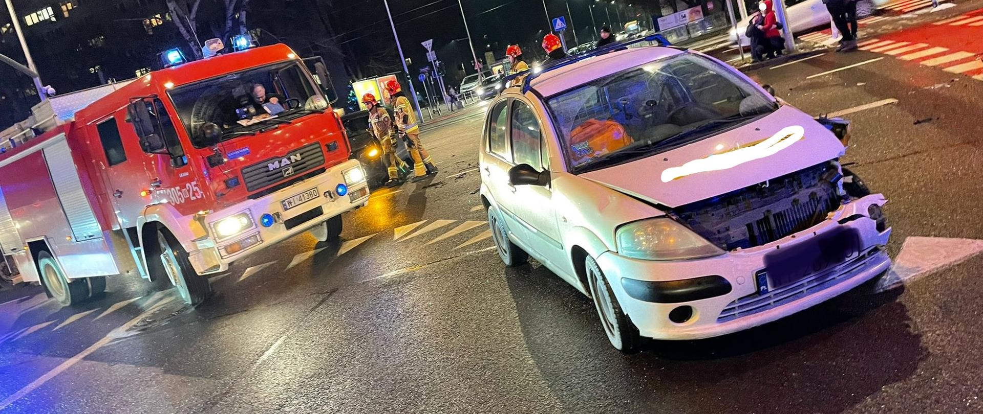 Na pierwszym planie widać rozbity biały samochód osobowy marki citroen w dalszym planie po lewej stronie widać wóz strażacki zabezpiecz jacy miejsce wypadku. 