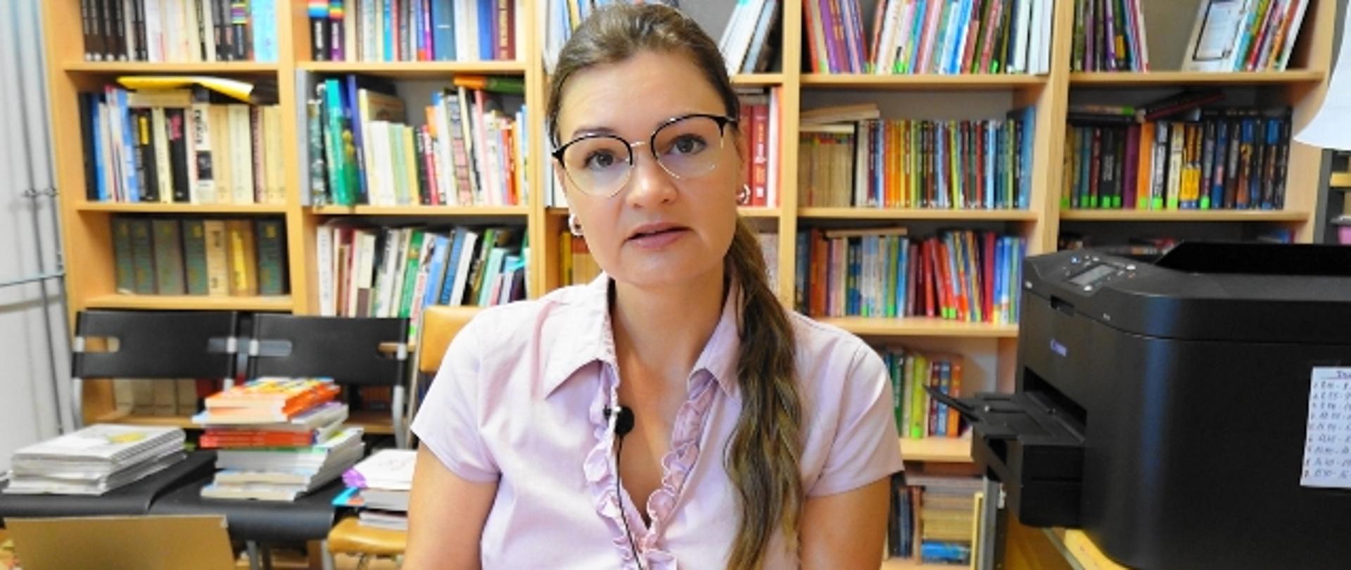 Nauczycielka trzyma książkę w ręku, w tle książki na półkach obok stoi drukarka