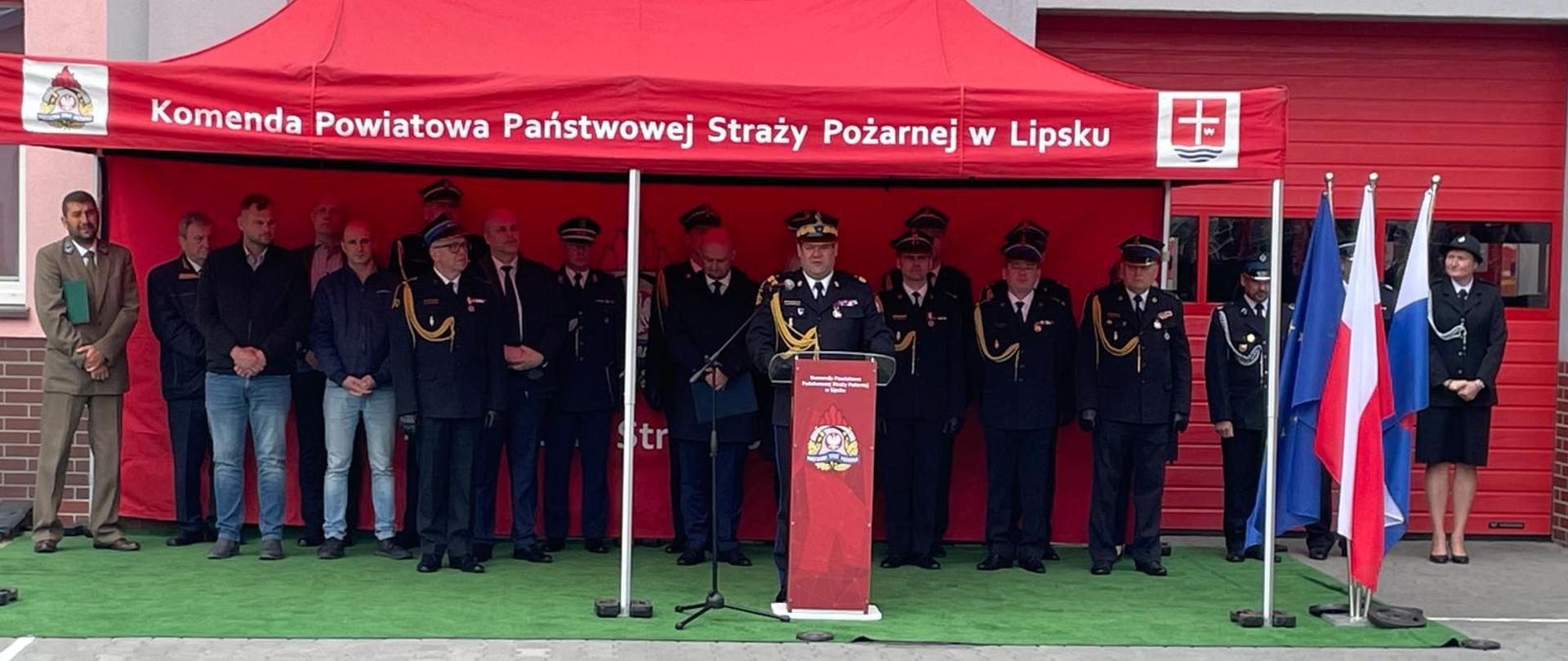 Funkcjonariusz Państwowej Straży Pożarnej w mundurze galowym przemawia zza czerwonej mównicy z godłem. Stoi pod dużym czerwonym namiotem z napisem „Komenda Powiatowa Państwowej Straży Pożarnej w Lipsku”. Za nim, w rzędzie, stoją inni strażacy oraz zaproszeni goście w cywilnych ubraniach. Po prawej stronie widoczne są flagi: Unii Europejskiej oraz dwie flagi Polski.