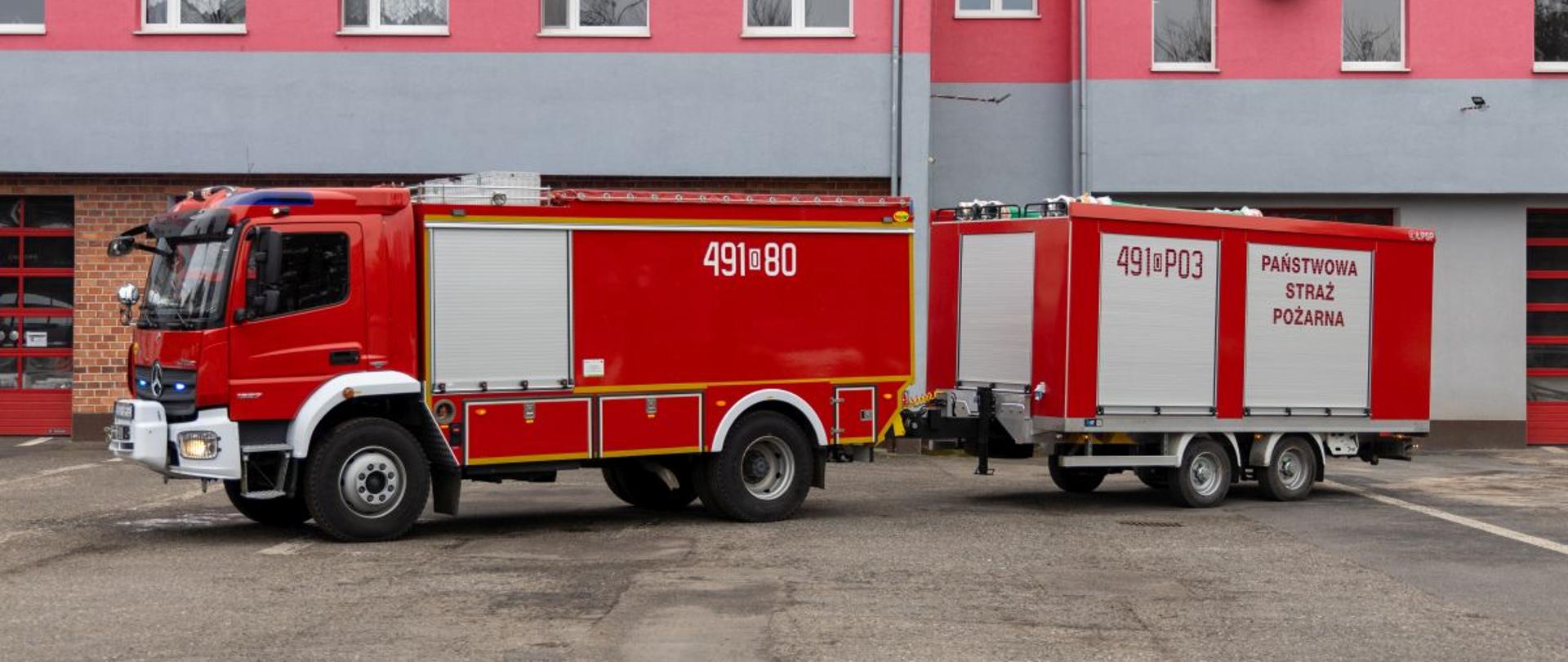 Czerwony samochód pożarniczy z oznaczeniem 491[O]80 stoi obok przyczepy 491[O]03 Państwowej Straży Pożarnej. W tle widoczny jest nowoczesny budynek strażnicy. Zestaw prezentuje mobilne wyposażenie jednostki PSP.