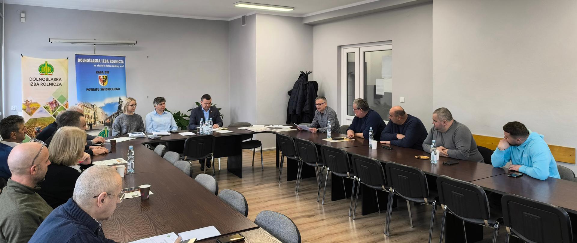 Grupa osób bierze udział w naradzie przy długich stołach ustawionych w sali o jasnych ścianach i drewnianej podłodze. Uczestnicy siedzą naprzeciw siebie. W tle widać drzwi, wieszak z płaszczami oraz plakaty informacyjne związane z Dolnośląską Izbą Rolniczą. 