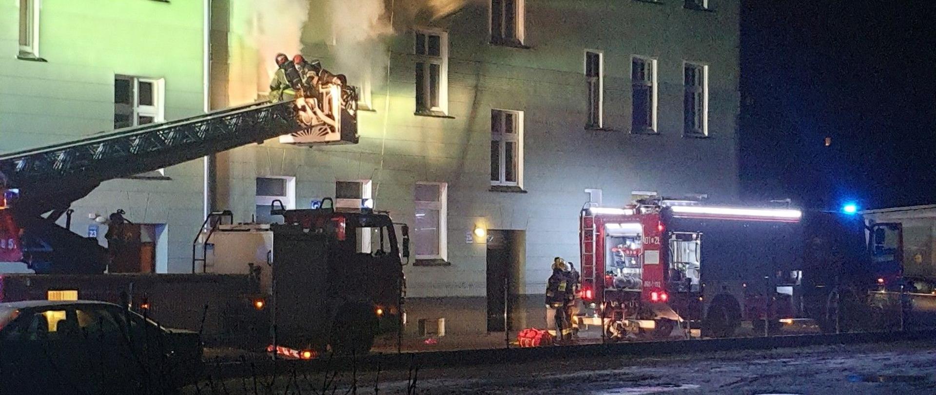Na zdjęciu widoczne dwa pojazdy pożarnicze w porze nocnej przy pożarze mieszkania w budynku wielorodzinnym w Brzegu. Z dwóch okien mieszkania na 1 piętrze wydobywa się gęsty dym.