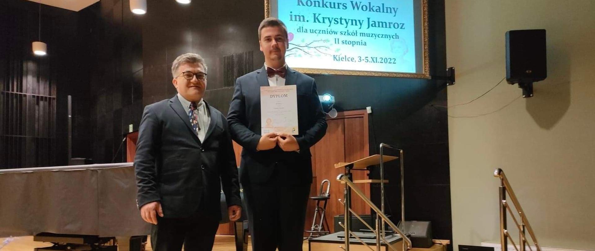zdjęcie przedstawia Szymona Maziarza trzymającego zdobyty dyplom i jego nauczyciela Marcina Pokusę na scenie w jednej z kieleckich szkół muzycznych z fortepianem i czarnym wykończeniem ścian