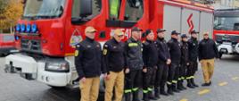 Strażacy stojący na baczność na tle samochodu pożarniczego.