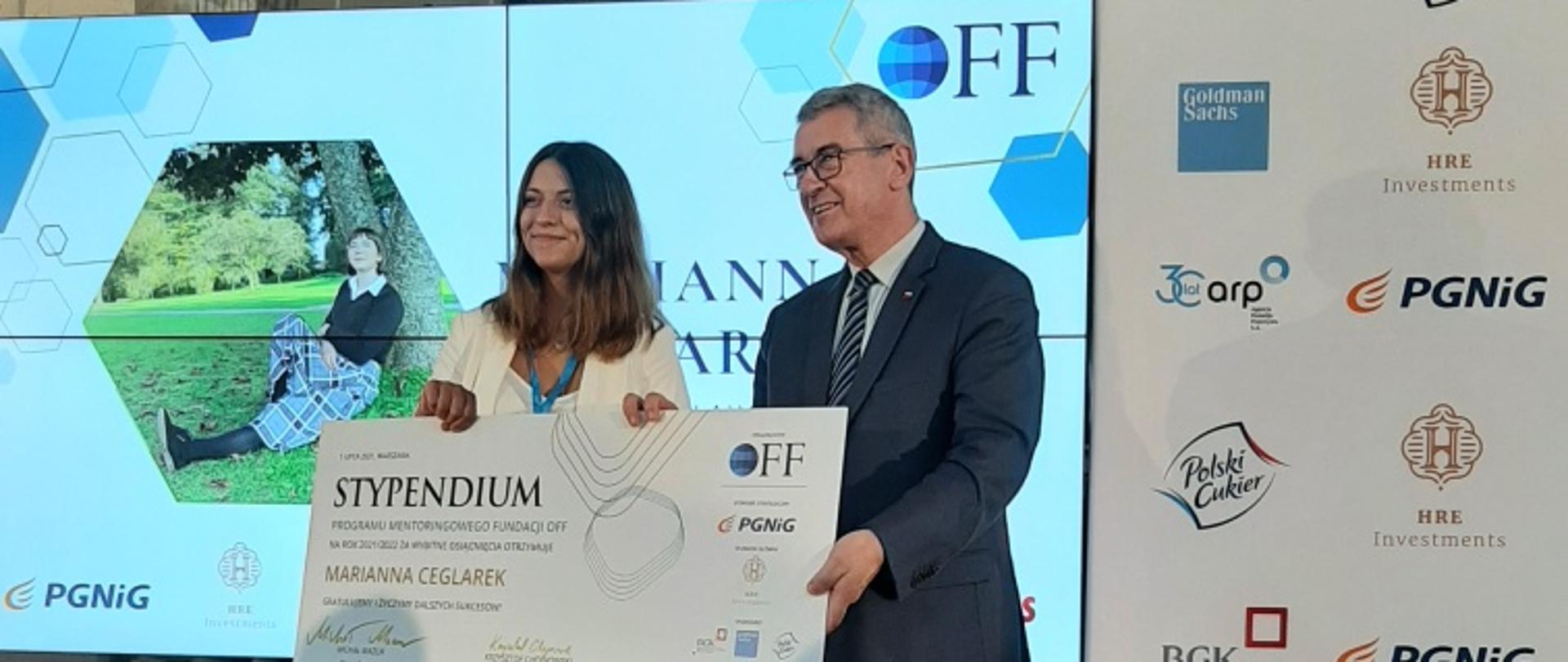 Wiceminister Wojciech Murdzek i laureatka stypendium programu Our Future Foundation. Kobieta jest ubrana w biały garnitur. Minister Wojciech Murdzek wręcza laureatce symboliczny dyplom stypendium. W tle ekran i baner z logotypami sponsorów. 