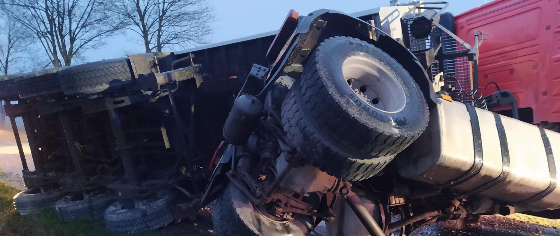 Naczepa typu wanna, leżąca na boku, częściowo na drodze częściowo w rowie, uszkodzony ciągnik siodłowy, pokrzywiona rama, ciągnik stoi na kołach