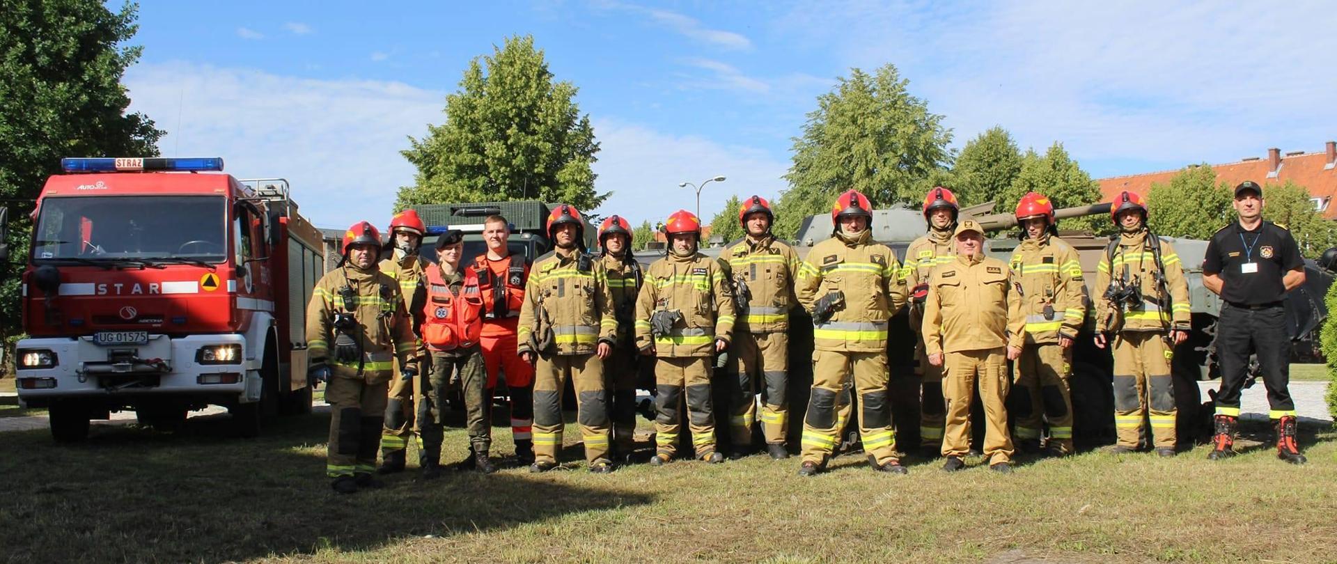 Na zdjęciu grupa strażaków w mundurach bojowych i czerwonych hełmach. Z lewej strony czerwony samochód strażacki. Niebieskie niebo, słonecznie.
