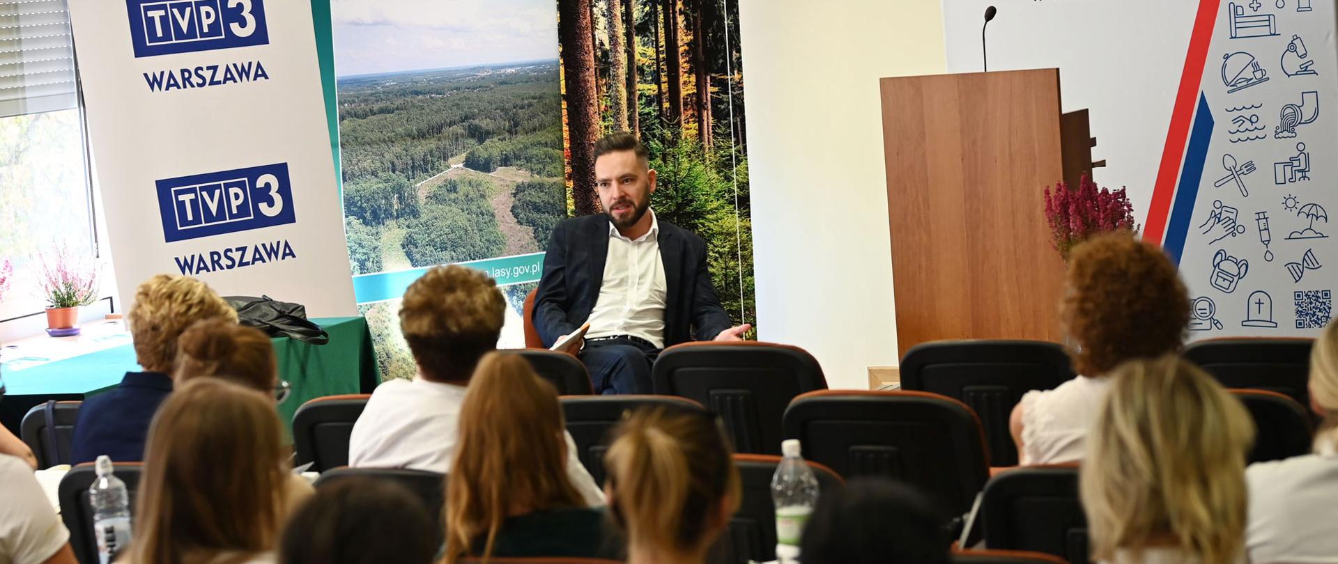 Mężczyzna siedzi w sali konferencyjnej na krześle trzymając w ręku książkę. Przed nim siedzą ludzie biorący udział w szkoleniu.