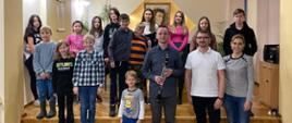 Kolorowa fotografia wykonana w auli PSM I st. w Kłodzku. Na zdjęciu widoczna grupa osób, tj. dr. Paweł Kroczek, który w ręku trzyma klarnet, obok nauczyciele Przemysław Polak i K. Brzeska Krawczyk za nimi grupa dzieci, które uczestniczyły w warsztatach klarnetowych. Zdjęcie wykonane 07.11.2022.
