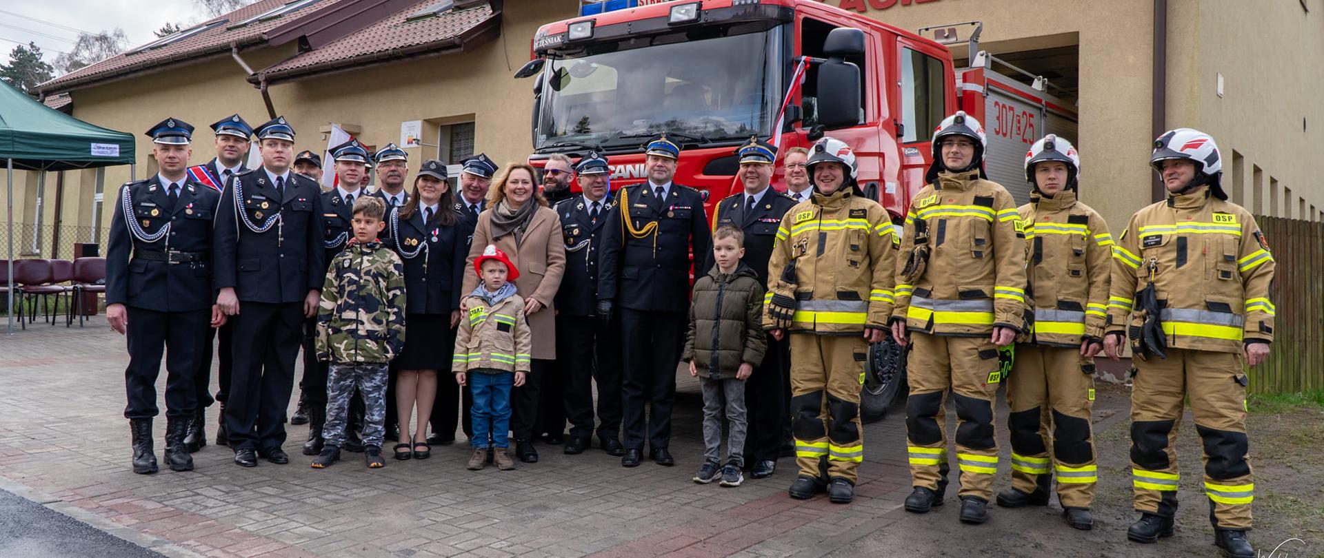 uczestnicy uroczystości, głownie druhowie i strażacy, pozują do zdjęcia na tle samochodu gaśniczego i remizy