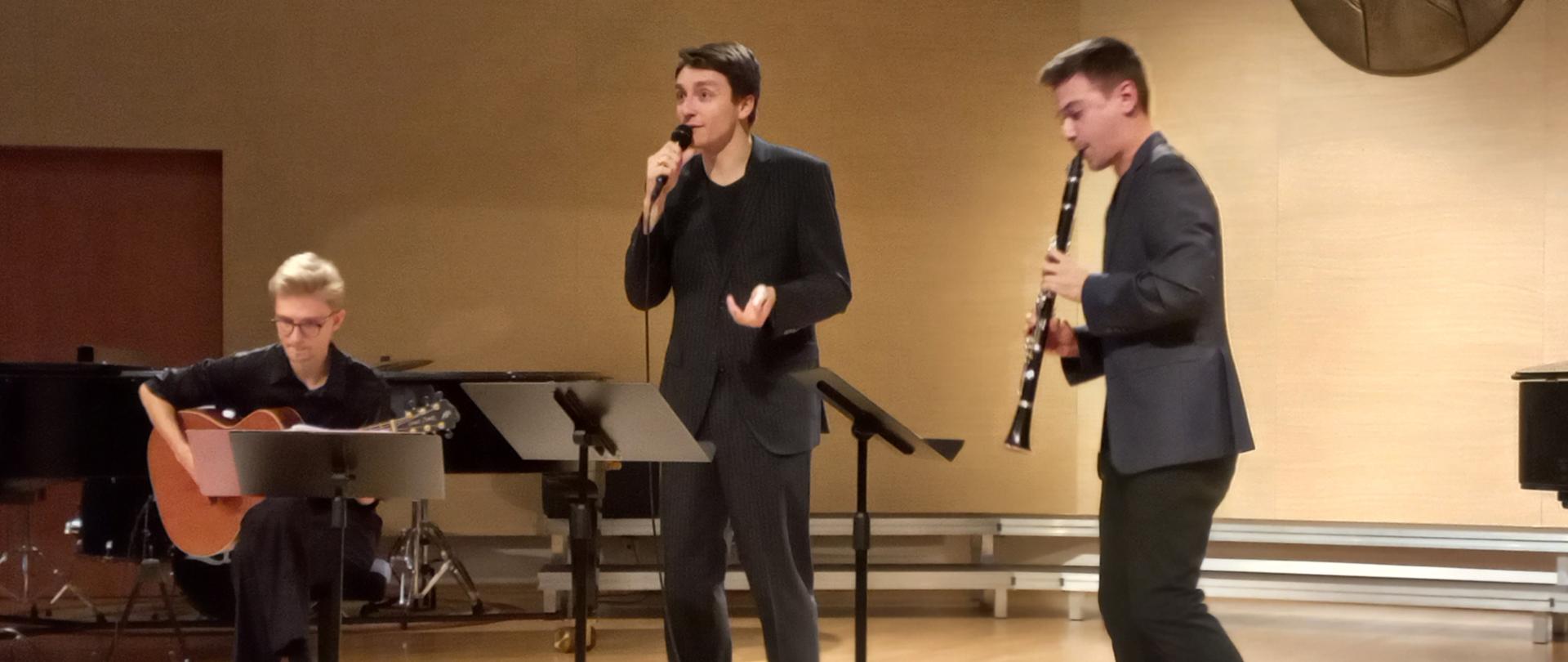Zdjęcie przedstawia trójkę artystów występujących na scenie sali koncertowej. Mężczyźni ubrani są w czarne garnitury. Dwóch mężczyzn gra na instrumentach i jeden śpiewa. Na scenie w tle znajdują się dwa fortepiany.