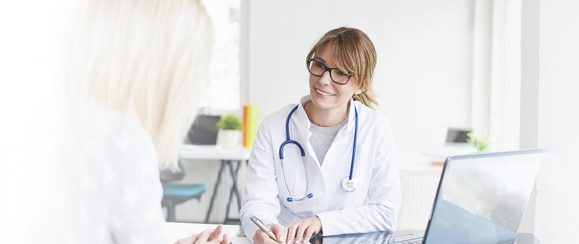 kobieta w białych fartuchu, z niebieskim stetoskopem na szyi, lekarz, rozmawia z pacjentem przy biurku, pacjentem jest kobieta