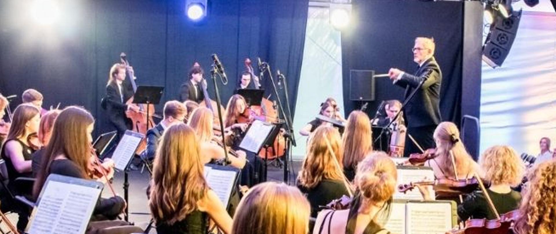 Czarne tło, w lewym górnym rogu zdjęcie orkiestry symfonicznej, informacje dotyczące koncertu uczniów ZSM w Bazylice Jasnogórskiej 14.12.2023 roku