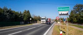 Na zdjęciu widać dwupasmową drogę prowadzącą do miejscowości Kolbuszowa. Po prawej stronie stoi zielona tablica z nazwą miasta. Wzdłuż drogi jadą samochody i ciężarówki, tworząc niewielki ruch. Po obu stronach znajdują się trawniki, chodnik oraz drzewa. Pogoda jest słoneczna, niebo jasne i bezchmurne.