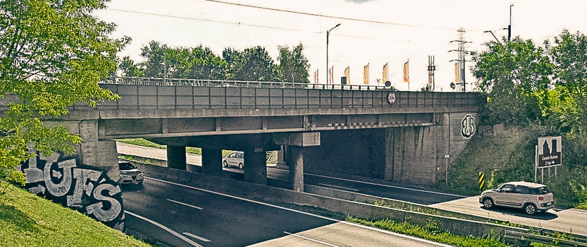 Widok na wiadukt tramwajowy nad DK86 w Będzinie. Dwuprzestrzenna droga krajowa przebiega w wykopie o mocno nachylonych skarpach. Na skarpach widoczna zieleń przydrożna. 