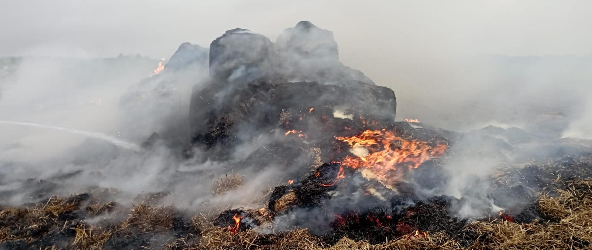 Zdjęcie przedstawia zgromadzone baloty słomy ogarnięte pożarem. Z lewej strony widać strumień wody podawany przez ratownika. 