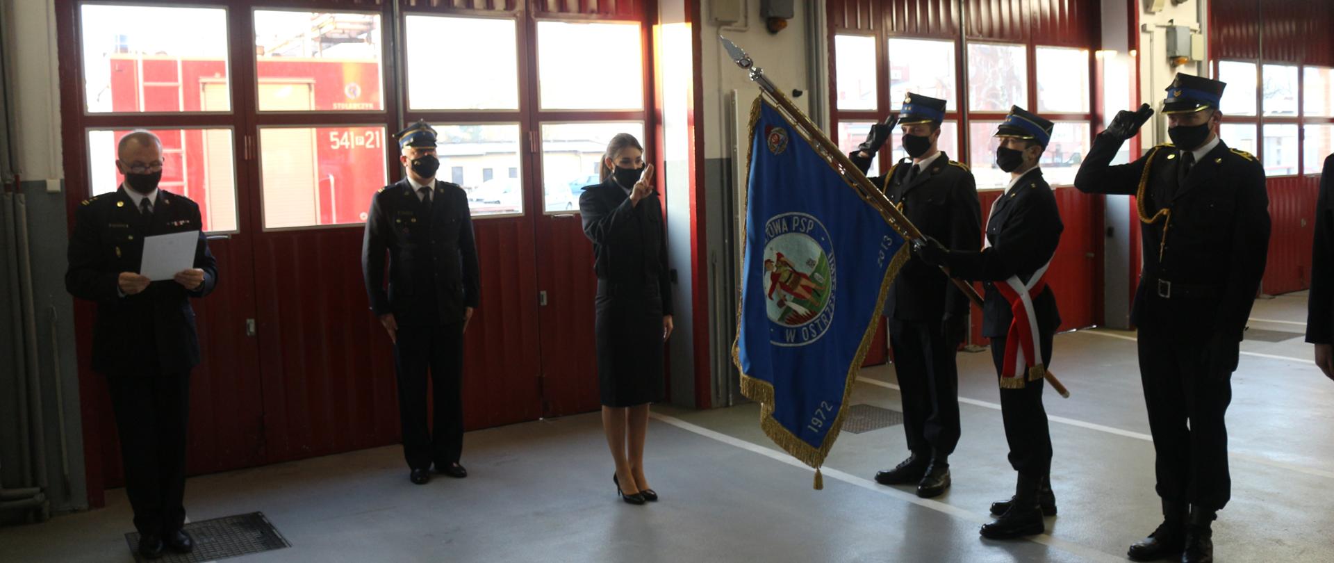 Zdjęcie przedstawia moment ślubowania nowego funkcjonariusza Państwowej Straży Pożarnej, który stoi na środku z podniesioną ręką. Po prawej stronie poczet sztandarowy z sztandarem w trakcie salutowania. Po lewej stronie Komendant Powiatowy PSP odczytuje rotę ślubowania. W tle widać samochód strażacki za bramami garażu.