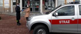 Strażak w czarnym mundurze przed budynkiem urzędu gminy, przekazuje ulotki kobiecie. Obok strażaka i kobiety samochód strażacki koloru srebrnego.