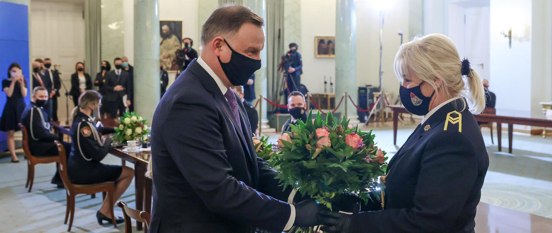 Kobieta ubrana w mundur Państwowej Straży Pożarnej odbiera z rąk mężczyzny ubranego w garnitur (prezydenta) bukiet kwiatów w tle stoły przy których siedzą inni strażacy oraz dziennikarze.