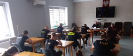 Zdjęcie przedstawia grupę ludzi siedzącą przy stolikach, ubranych w czarne umundurowanie koszarowe strażaka. Zdjęcie zrobiono podczas pisania egzaminu.