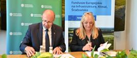 Na zdjęciu na tle banera z logo Narodowego Funduszu Ochrony Środowiska i Gospodarki Wodnej siedzą przy stole od lewej Zastępca Prezesa Narodowego Funduszu Ochrony Środowiska i Gospodarki Wodnej Robert Gajda i prezes Przedsiębiorstwo Wodociągów i Kanalizacji "WODNIK" Katarzyna Wierska-Kuberska. Zdjęcie przedstawia moment podpisania umowy. 