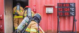 Strażacy trzymając wąż gaśniczy wchodzą do komory celem przeprowadzenia ćwiczenia gaśniczego.
