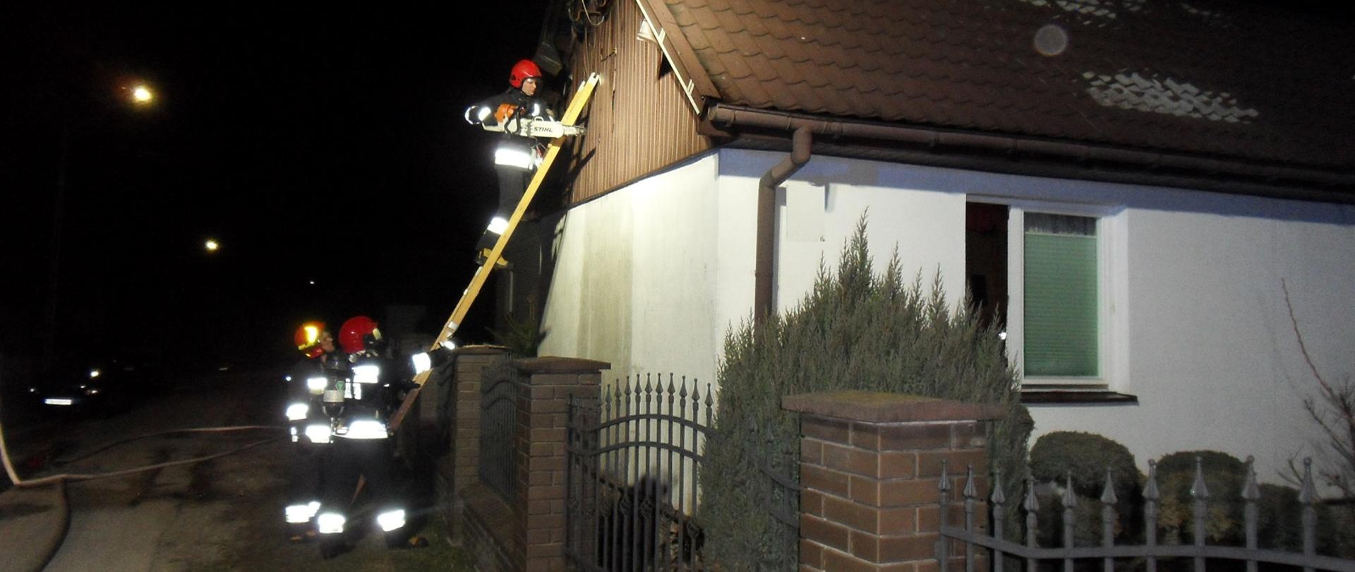 Zdjęcie przedstawia strażaków podczas działań ratowniczo gaśniczych. Dwóch z nich podtrzymuje drabinę, na której działania prowadzi kolejny strażak. Drabina oparta o budynek mieszkalny.